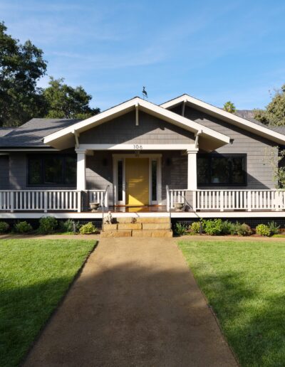 A single-story gray house with a yellow door, white trim, and a large front porch, surrounded by a well-kept lawn and trees.