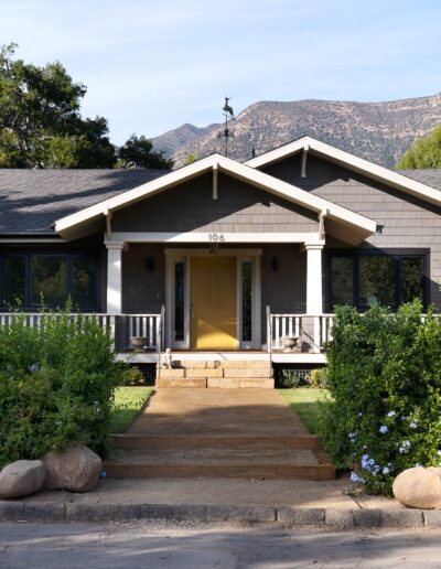 A single-story house with a gray exterior, yellow front door, and surrounding greenery. Steps lead up to the entrance, and mountains are visible in the background.