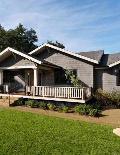 A single-story house with gray siding and a front porch, surrounded by a well-kept lawn and trees, under a clear sky.