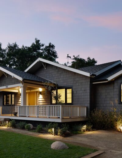 Single-story house with dark gray siding and a large wooden porch, set in a landscaped yard at dusk.