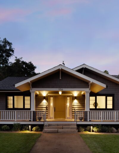 A single-story house with a gray exterior and white trim is lit up at dusk. It has a welcoming front porch, neatly trimmed lawn, and trees in the background.