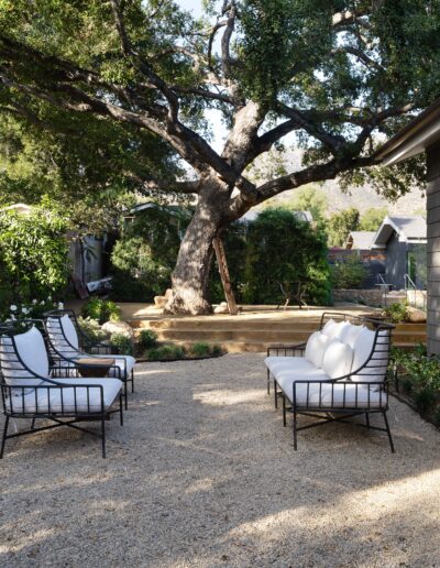 A serene backyard patio with four cushioned chairs, gravel ground, and a large tree providing shade. Lush greenery and white flowers border the area next to a dark-colored house.