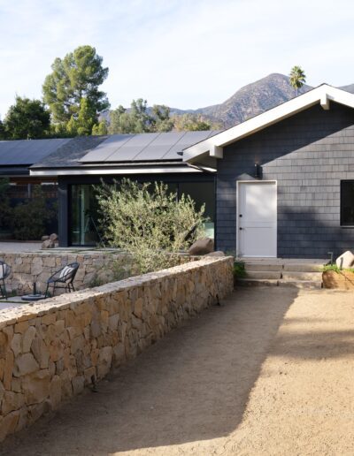 A single-story house with solar panels on the roof, a stone wall around a small pool area, and mountain scenery in the background, surrounded by greenery.