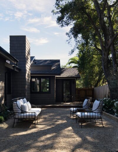 A backyard with gravel ground, black-painted house exterior, cushioned metal chairs, and well-maintained vegetation with trees and shrubs.