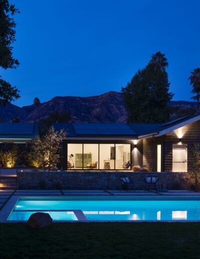 Modern house with large windows illuminated at twilight, surrounded by trees and mountains, featuring a lit swimming pool in the foreground and a neatly landscaped yard.