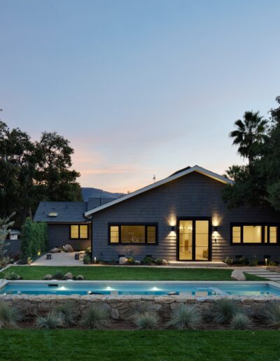 A modern single-story house with large windows is lit up at dusk, featuring a backyard with a swimming pool, stone landscaping, and surrounded by trees.