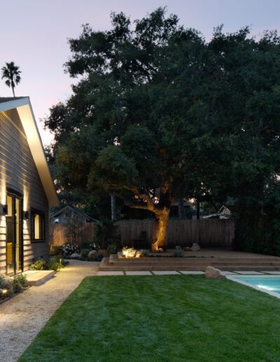 A modern house with a lawn, lit pathway, and swimming pool surrounded by trees in the backyard during dusk.