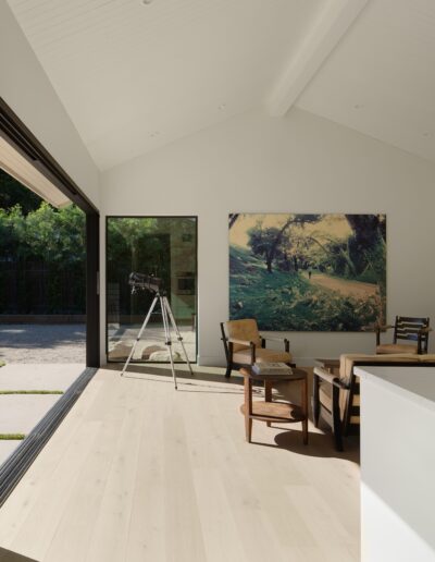 A modern, minimalist room with large sliding glass doors, a telescope by the door, a painting on the wall, wooden furniture, and a small potted plant on a white counter.