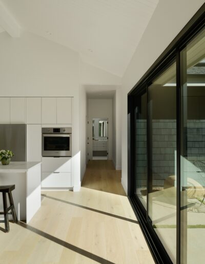 Modern kitchen with white cabinets and countertops, an island with two black stools, an open sliding glass door leading to a shaded outdoor area with a lawn and trees. Natural light fills the space.