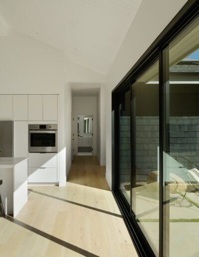 Modern kitchen with white cabinets, a marble backsplash, and an island with two black stools. Large sliding glass doors open to an outdoor patio with lounge chairs and greenery.