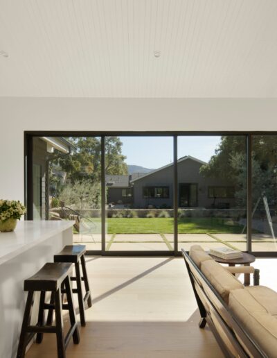 A modern kitchen and living area with white cabinetry, a marble countertop, bar stools, a beige sofa, and a telescope by large glass doors overlooking a green backyard.