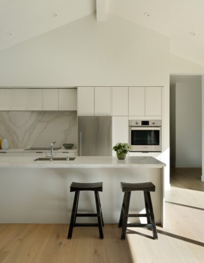 Modern kitchen with white cabinets, marble backsplash, stainless steel appliances, and a large island with two wooden stools, illuminated by natural light from a large window.