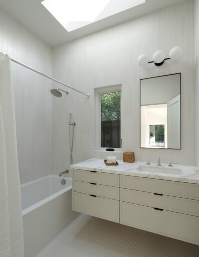 A modern, minimalist bathroom with white tiles, a bathtub, shower, dual sinks, a large mirror, and a window. Natural light enters through a skylight, illuminating the space.