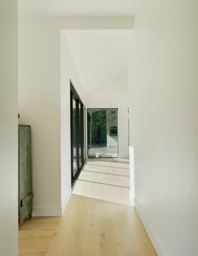 A hallway with light wooden flooring and white walls leads to a room with large floor-to-ceiling windows and sliding glass doors, providing a view of a bright outdoor space. An antique cabinet is on the left.