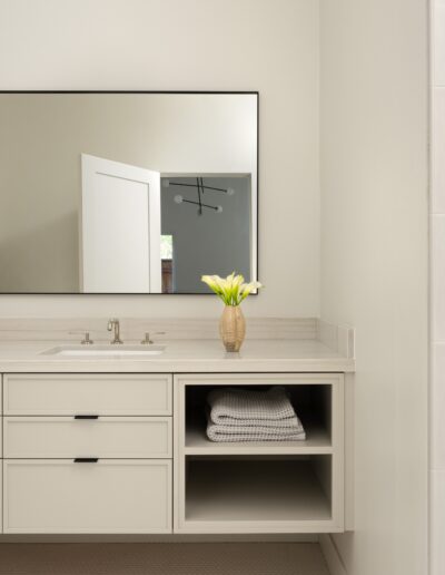 A modern bathroom with a large mirror, a countertop with a sink, storage drawers, a vase with flowers, neatly folded towels, and a partially visible shower area.