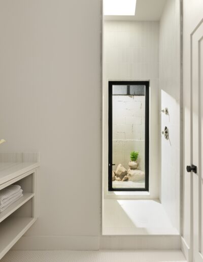 A minimalistic bathroom with white cabinetry, a wall-mounted mirror, a small vase with flowers, and an open shower area with a glass door leading outside to a small plant.