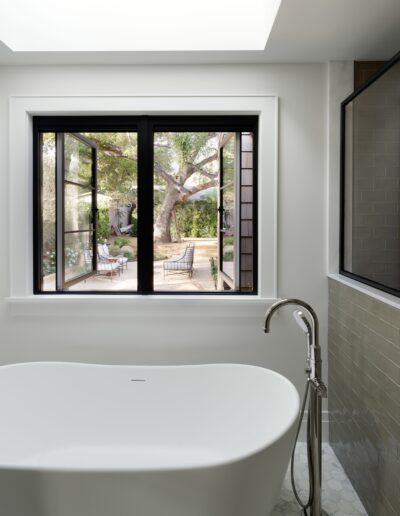 A white bathtub is positioned beneath an open black-framed window, offering a view of a backyard with an outdoor seating area and large trees.