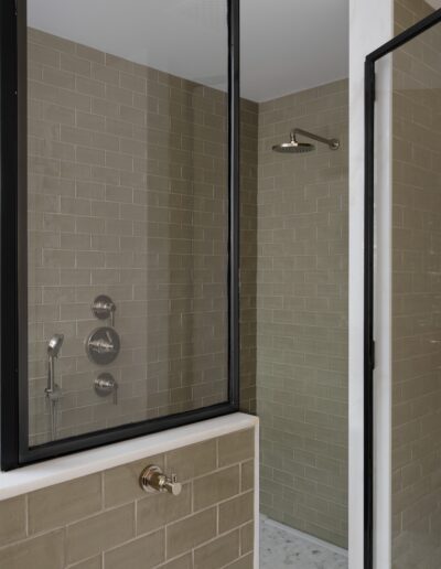 A glass-enclosed shower with beige subway tile walls and a black metal frame, featuring a showerhead, control knob, and handheld sprayer.