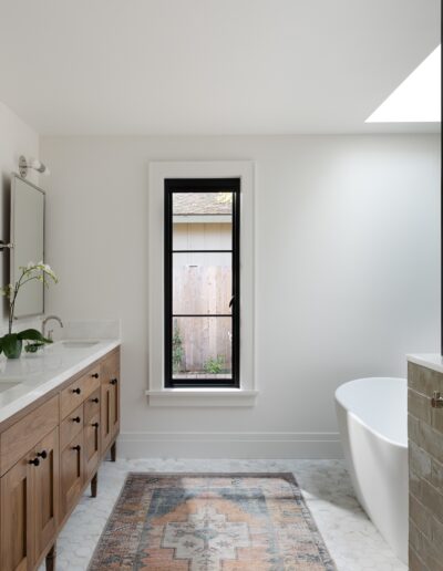 A modern bathroom with a double sink vanity, a freestanding bathtub, wooden cabinetry, a glass window, and a patterned rug on the floor.