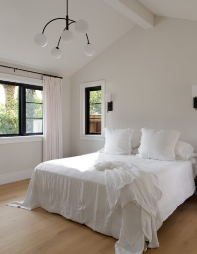 A minimalist bedroom with a white bed, pillows, a black nightstand, two windows with white curtains, and modern hanging light fixtures. The room has light wooden flooring and white walls.