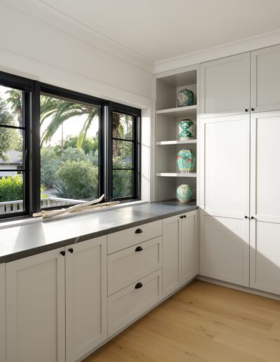 A bright kitchen with white cabinets, black window frames, and wooden flooring. Shelves with decorative vases are on the right side, while windows on the left offer a view of palm trees outside.