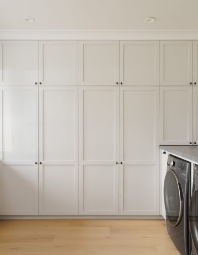 A modern laundry room with light gray cabinets, open shelving with decorative items, a gray countertop, and a matching washer and dryer set. Natural light streams in through a window on the left.