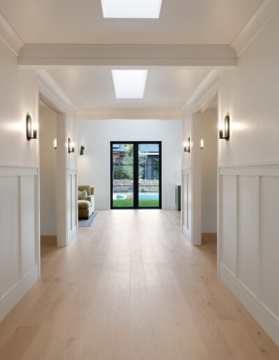 A long, empty corridor with white walls, wooden floor, and wall-mounted lights leading to a glass door overlooking a greenery-filled patio. Skylights provide additional natural light.