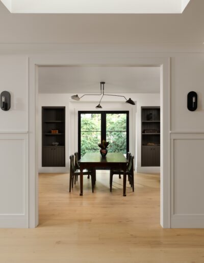 A dining room with a rectangular table and four chairs, lit by a ceiling light fixture. The room features two dark built-in cabinets and large windows with views of greenery.