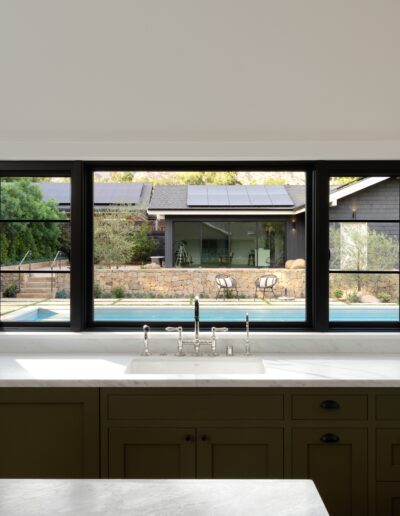 A kitchen window view showing a sink and faucet with a stone countertop and green cabinetry. Outside, a pool and two lounge chairs are visible, bordered by landscaping and a house.