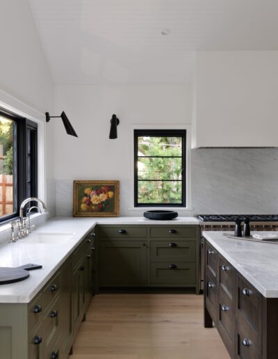 Modern kitchen with green cabinets, white countertops, black fixtures, and a black window. There is a painting on the countertop and a view of outdoor greenery through the window.