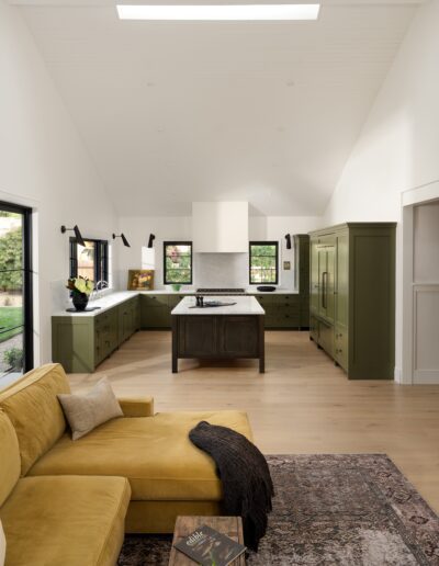 A modern kitchen with olive green cabinets, a large island with a white range hood, and a skylight. The space includes a yellow sofa, a patterned rug, and natural light from windows and doors.