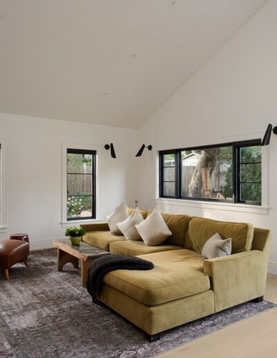 A modern living room with a yellow sectional sofa, a brown leather armchair, and a wooden coffee table. The room has white walls, black-framed windows, and wood flooring.