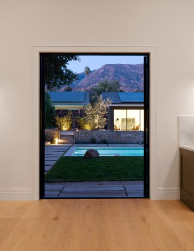 A modern living room opens onto a backyard with a swimming pool, green lawn, and mountain views in the background, framed by an open sliding door.