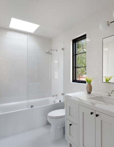 A modern white bathroom features a bathtub with a glass shower partition, a toilet, a marble countertop with a sink, and a large mirror above the vanity, illuminated by natural light from a skylight.