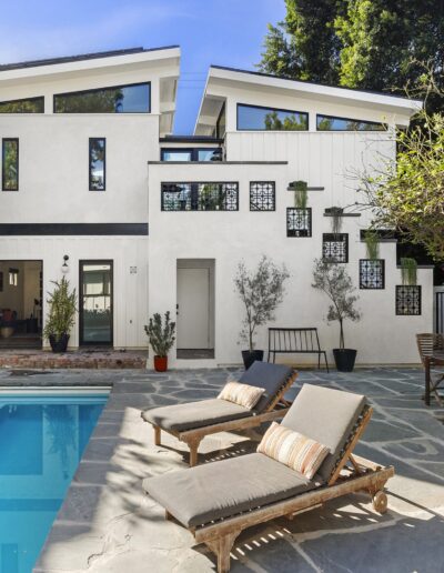 Modern two-story house with a white exterior, adjacent pool, patio, outdoor seating, and greenery. The patio has lounge chairs, a small table, and potted plants.