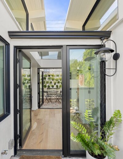 A modern glass door opens into a bright room with large windows and potted plants. A black outdoor light fixture is mounted on the white exterior wall, and a mat is placed at the entrance.