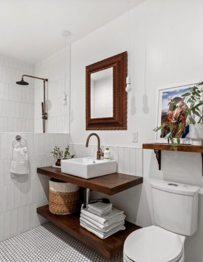 A modern bathroom with a walk-in shower, wall-mounted sink, mirror, toilet, and wooden shelving with plants and a framed picture. The design includes white tiles and a mix of dark wood elements.