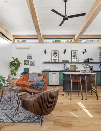 A modern open-concept living space with a kitchen, sitting area, and dining area. Features light wood floors, green cabinets, a ceiling fan, and large windows. Furnished with plants, chairs, and a rug.