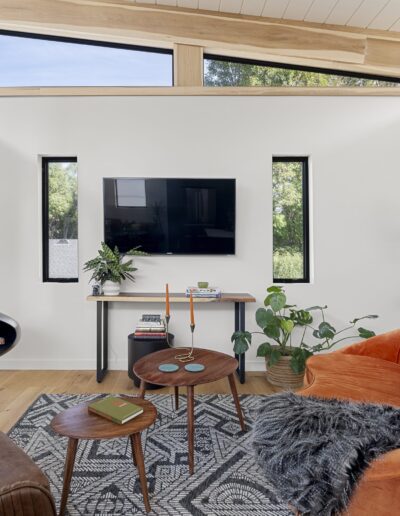 A modern living room with large windows, a wall-mounted TV, a hanging fireplace, and a mix of orange and brown seating. The room has green plants, a patterned rug, and a small wooden table.