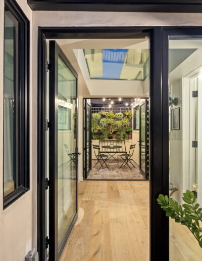 View through black-framed glass doors into a bright hallway with a wooden floor, leading to a patio with a table, chairs, and wall-mounted plants.