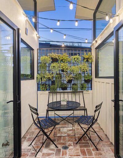 A narrow patio with glass doors, a brick floor, a small black metal table, two chairs, string lights hanging above, and a plant wall in the background.