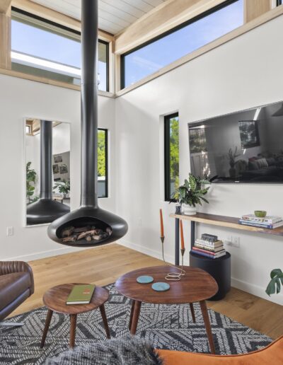 A modern living room featuring a wall-mounted TV, a suspended fireplace, a leather armchair, a coffee table with books and a laptop, and multiple windows allowing natural light into the space.