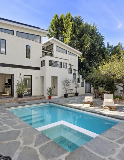 A modern two-story house with a swimming pool in the backyard, surrounded by outdoor furniture and greenery, under a clear sky.