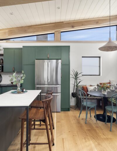 Modern kitchen with green cabinets, an island with stools, stainless steel appliances, and a cozy dining nook with green seating and pillows against the wall. Plentiful natural light from large windows.