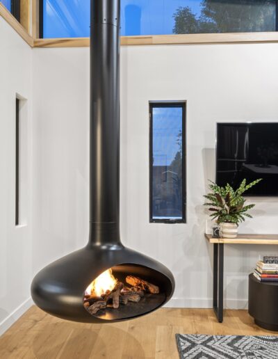 Modern living room with a suspended black fireplace, wooden flooring, and a wall-mounted TV. A potted plant sits on a table beneath the TV, and a rug covers part of the floor.