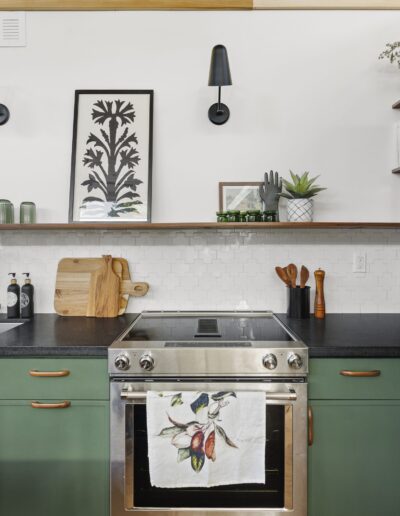 A modern kitchen with green cabinets, black countertop, stainless steel stove, and a framed botanical artwork above the backsplash. A towel with fruit prints hangs on the oven door.