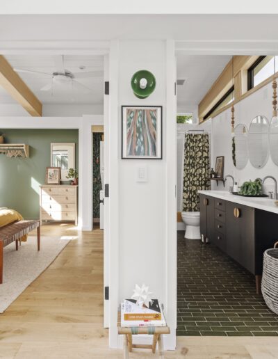 A hallway leads to a bedroom on the left and a bathroom on the right. The bathroom has double sinks and a green tile floor, while the bedroom features a wooden dresser and a green accent wall.