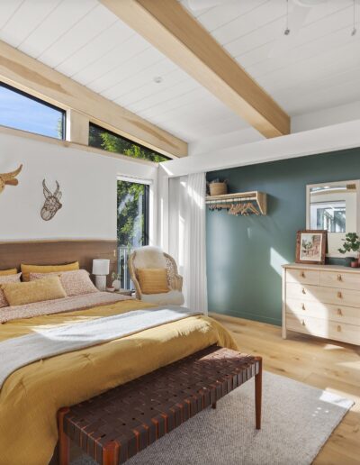 A modern bedroom with a wooden bed, yellow bedding, large window, green accent wall, dresser, and open closet. Sunlight illuminates the room. Ceiling beams and nature-themed decor are visible.