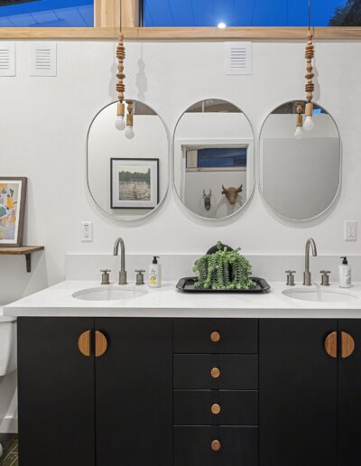 Modern bathroom with double sink vanity, black cabinets, round mirrors, decorative plants, and picture frames. Shower has a patterned curtain. Hanging lights and wicker storage basket are present.