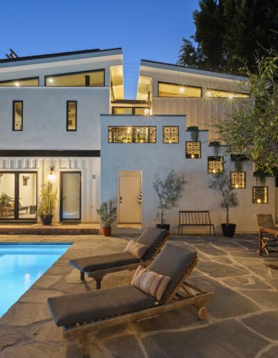 Modern two-story house with white exterior, large windows, outdoor pool, lounge chairs, patio furniture, and surrounding plants, viewed at dusk.